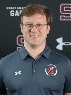 David W. smiling while wearing a gray polo shirt in front of a dark athletic backdrop.