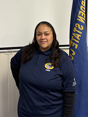 Myrisila V. smiling indoors while wearing a Garden State Council staff shirt, with a council flag beside her.