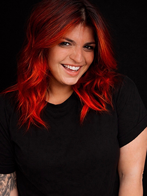 Priscilla B. smiling against a dark background with bright red hair.