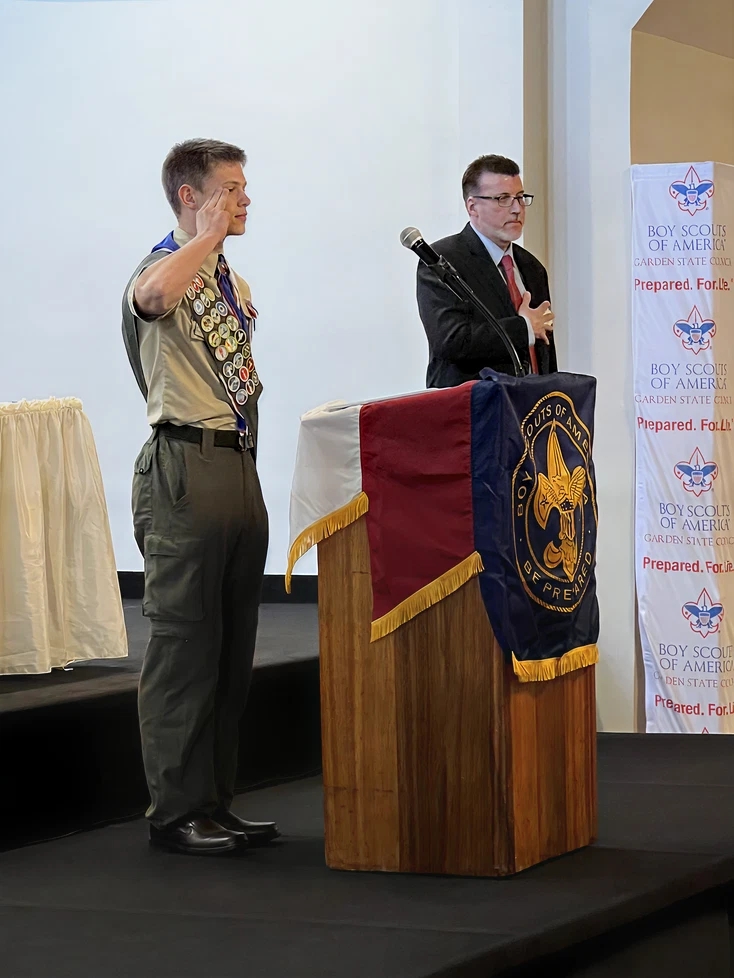 Eagle Scout raising his hand during a ceremony beside an adult standing at a podium.