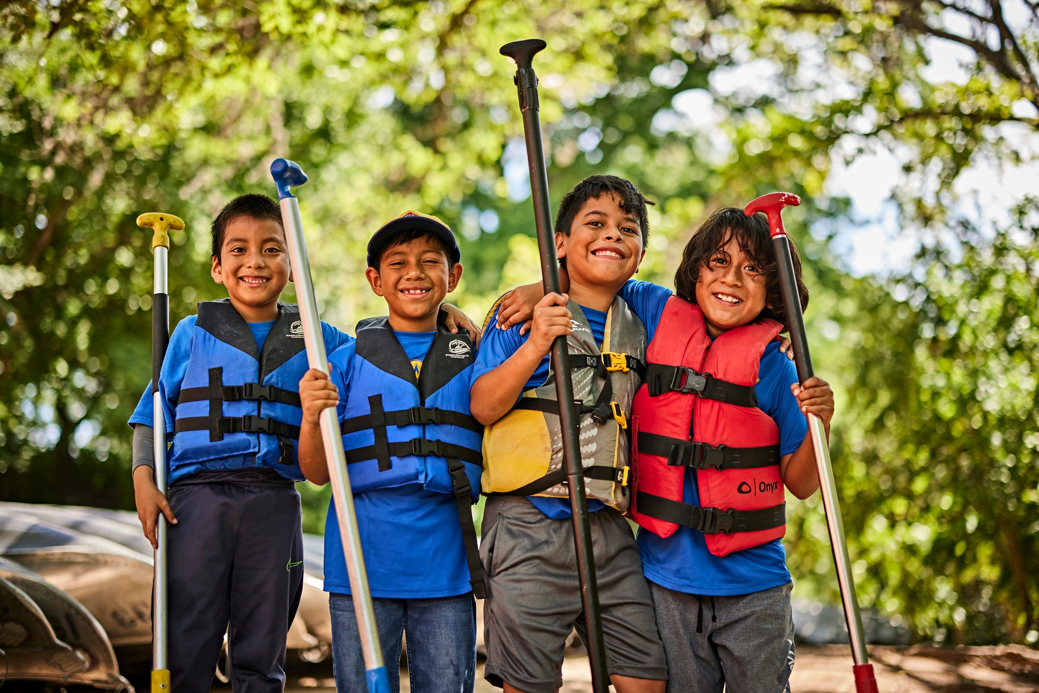 Four smiling Scouts wearing life jackets and holding paddles outdoors near the water.