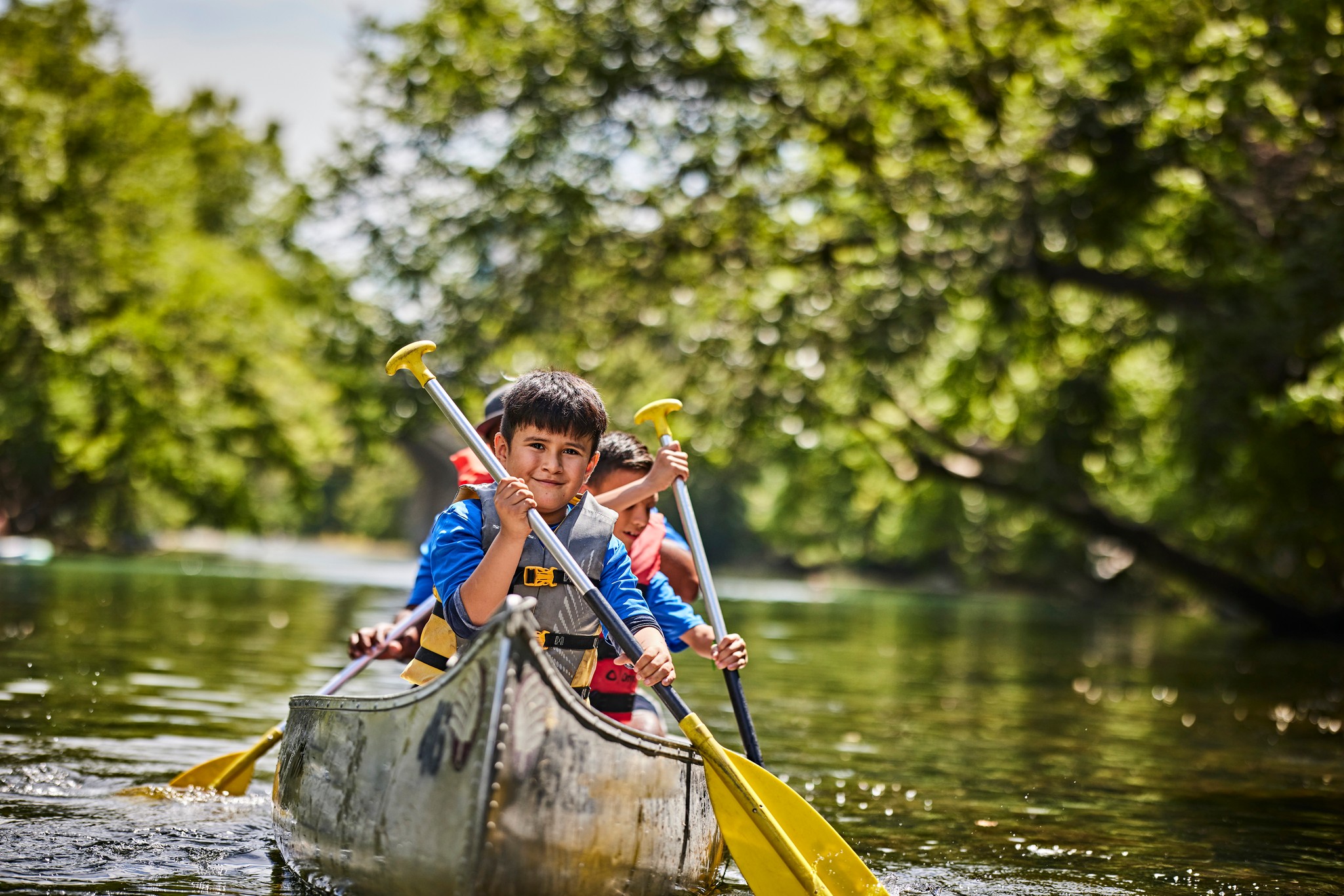 Scouts wearing life jackets paddling a canoe together on a tree-lined lake.