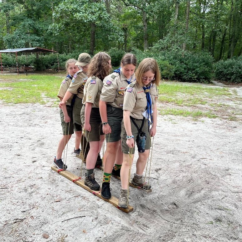 Five Scouts working together to walk on wooden planks during an outdoor team-building activity.
