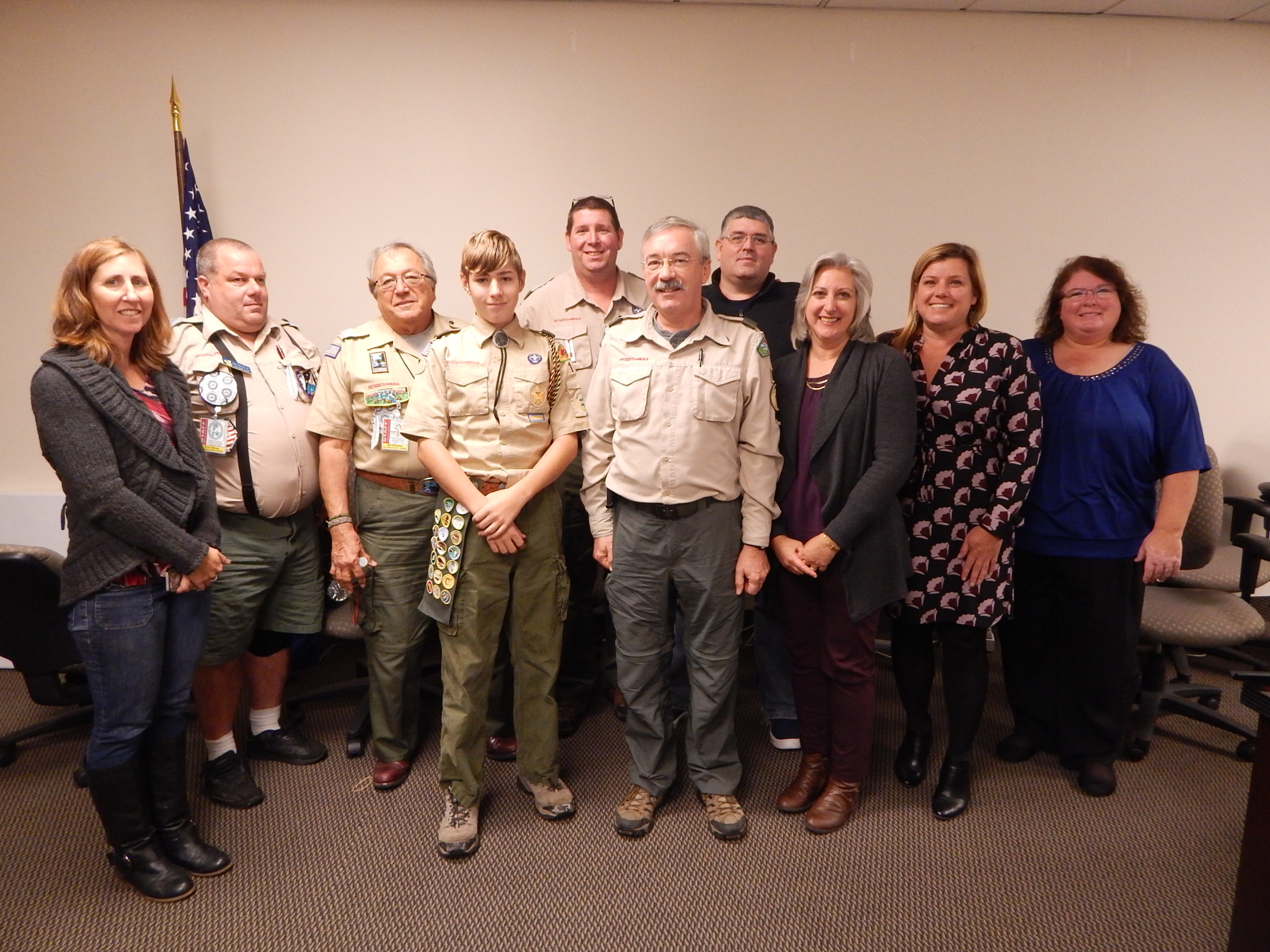 Scout in uniform standing with adult Scouting leaders and community members during an indoor recognition gathering.