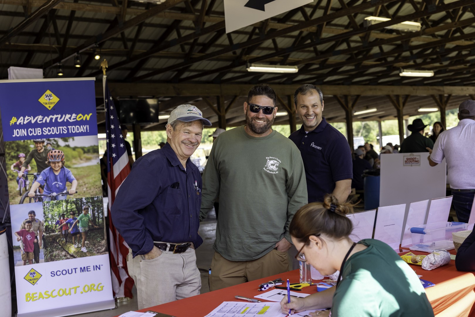 Three men posing at a community event beside a Cub Scouts recruitment display.