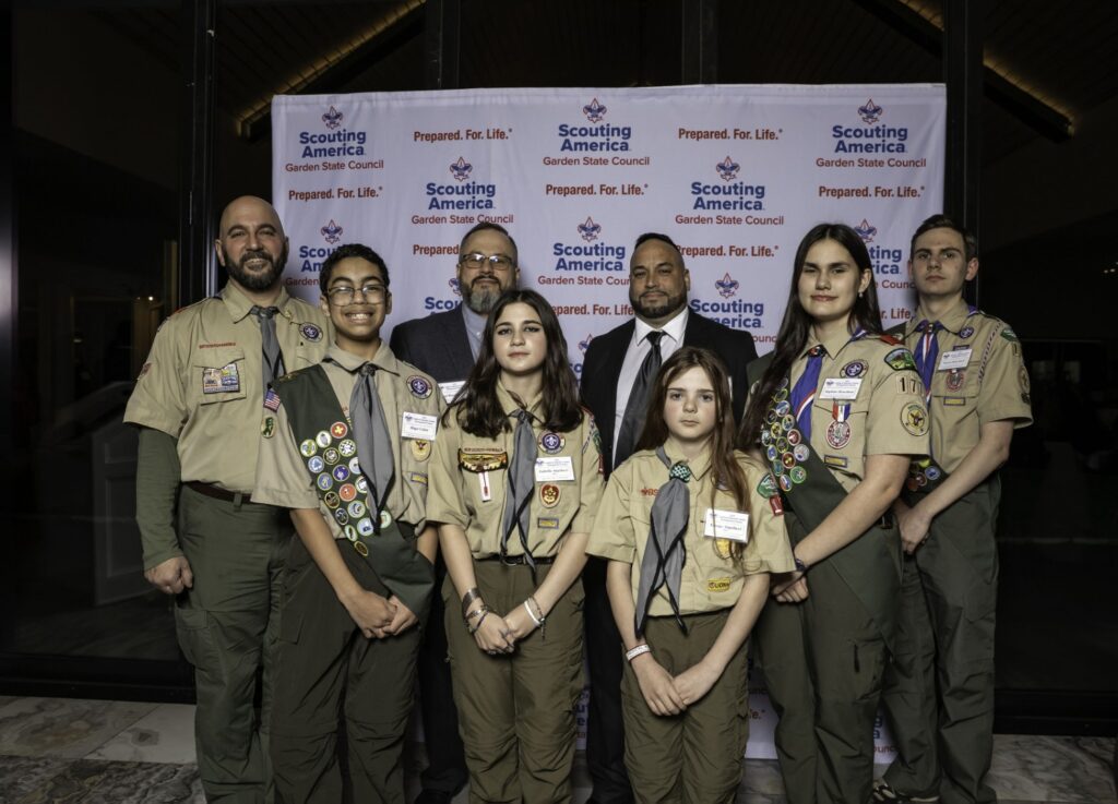 Scouts and adult leaders posing together in front of a Scouting America Garden State Council backdrop.