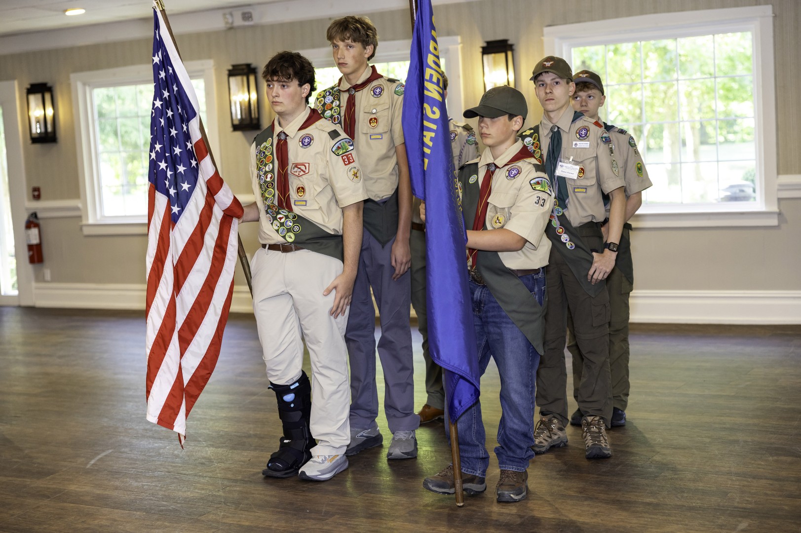 Scouts in uniform presenting the American flag and a troop flag during an indoor color guard ceremony.