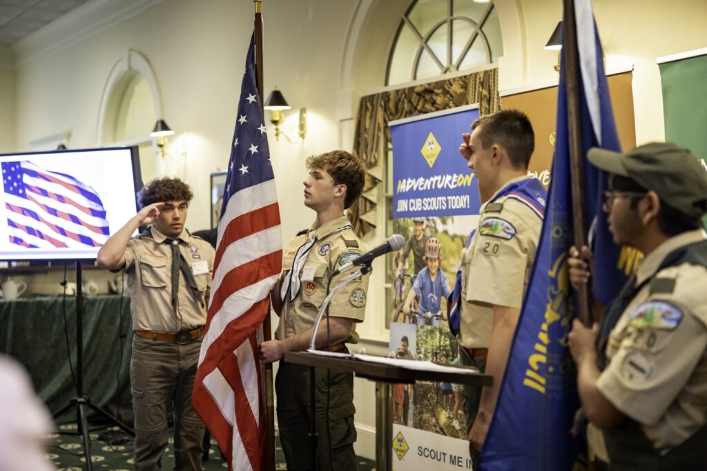 Scouts in uniform presenting the American and council flags during an indoor ceremony.