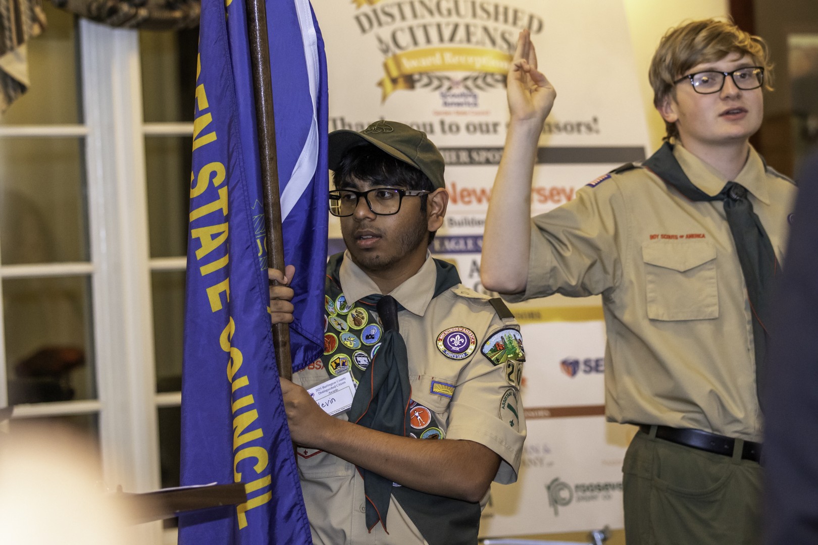 Two Scouts participating in a ceremony at a Distinguished Citizens event, with one holding a Garden State Council flag.