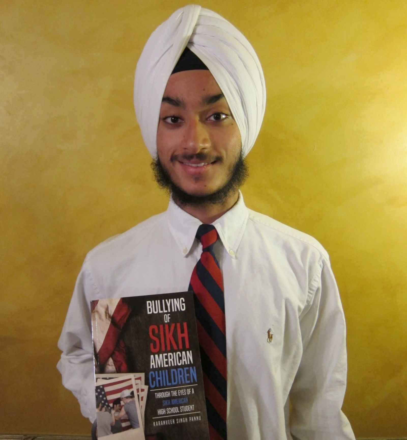 Karanveer Singh Pannu, a Sikh-American student holding the book Bullying of Sikh American Children while wearing a white turban and striped tie.