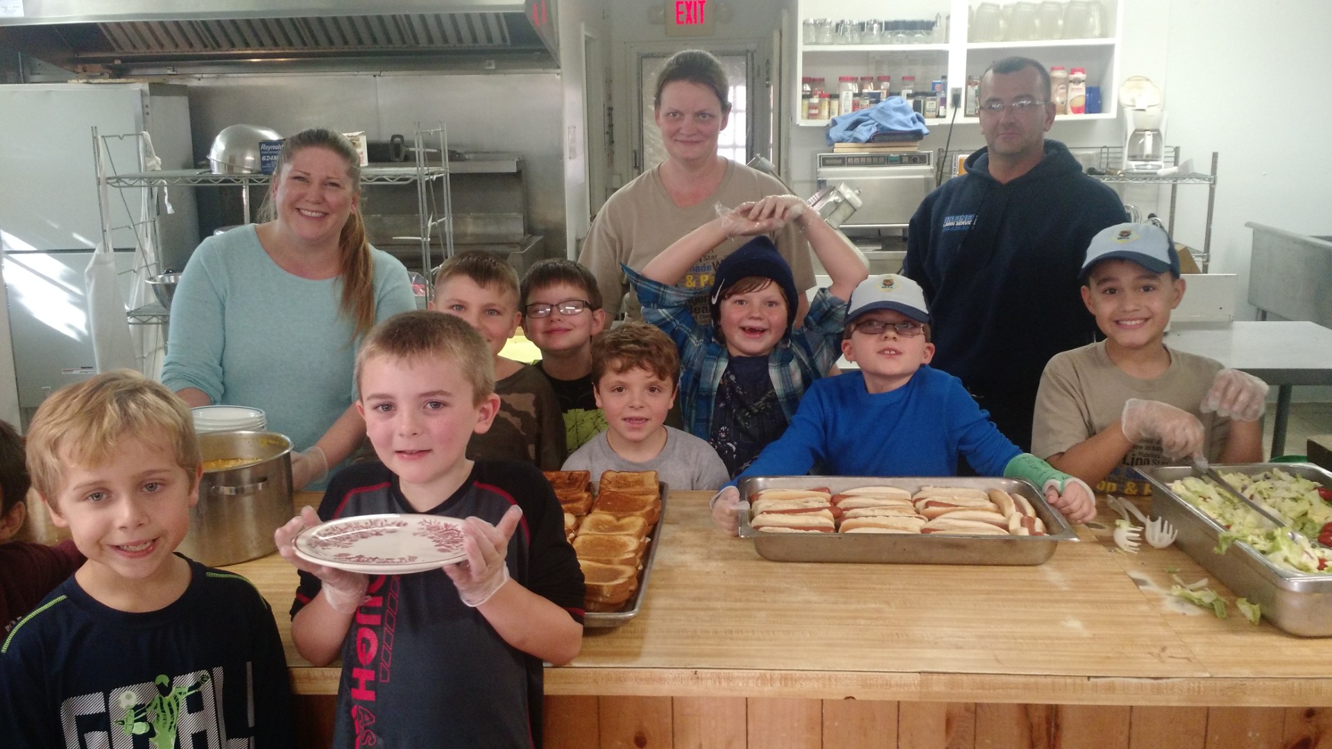 Young Scouts and adult volunteers preparing sandwiches and salad together in a camp kitchen.