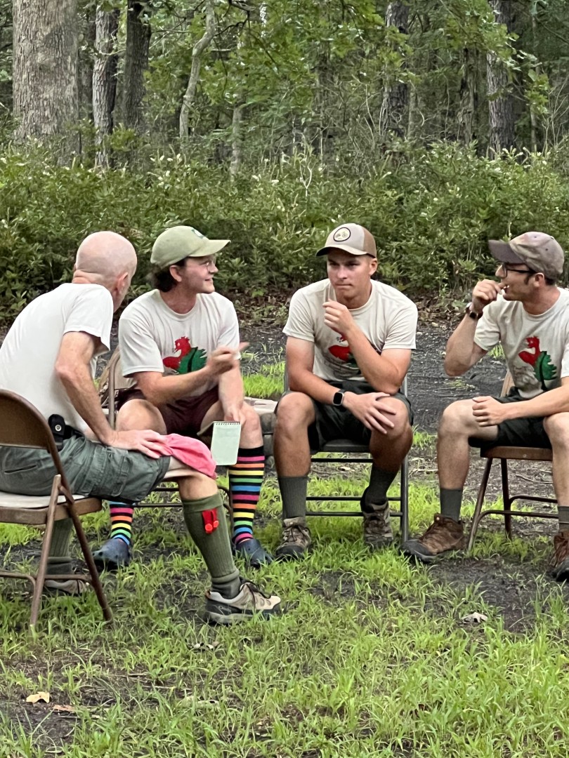 Four adult Scouting leaders sitting in a circle and talking outdoors during 2023 Trek Camp.