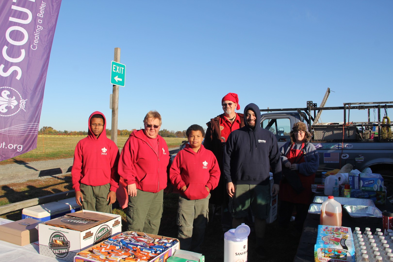 Scouts and adult volunteers standing behind a refreshment table at an outdoor Scouting event.