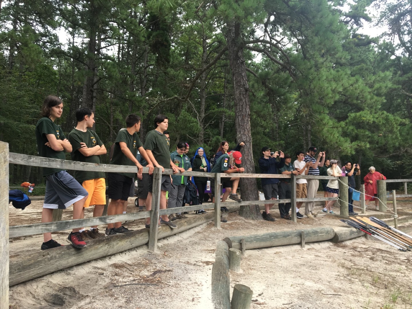 Scouts and visitors gathered along a wooden railing to watch an outdoor activity at camp.