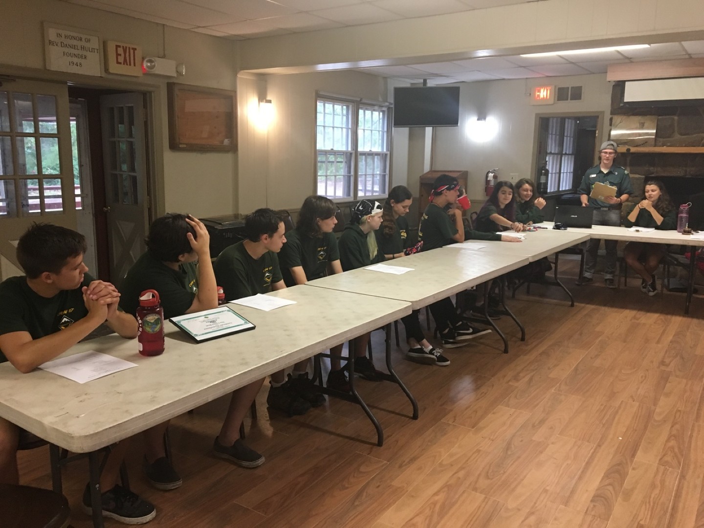 Scouts seated around tables during an indoor group meeting at camp.