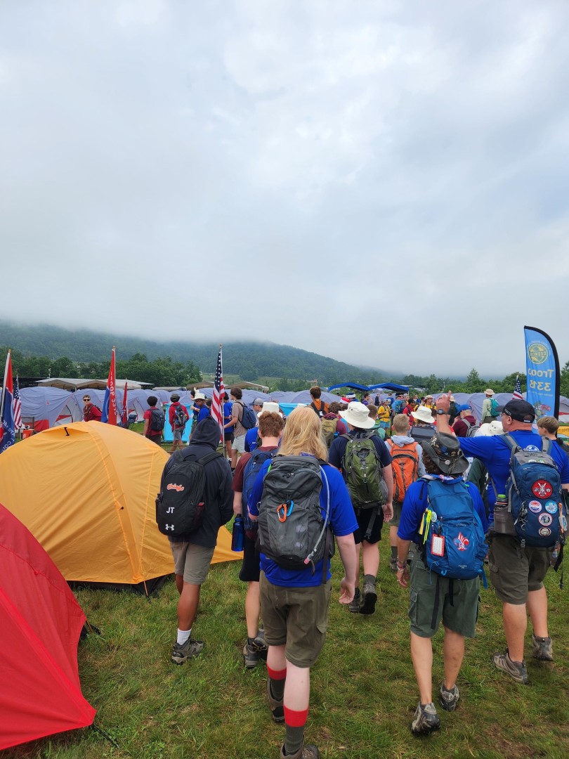 Scouts carrying backpacks walking through a large tent campsite during a Scouting jamboree.