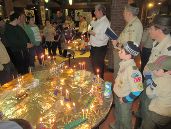Scouts and adult leaders gathered around a large display of lit menorahs during a Chanukah celebration.
