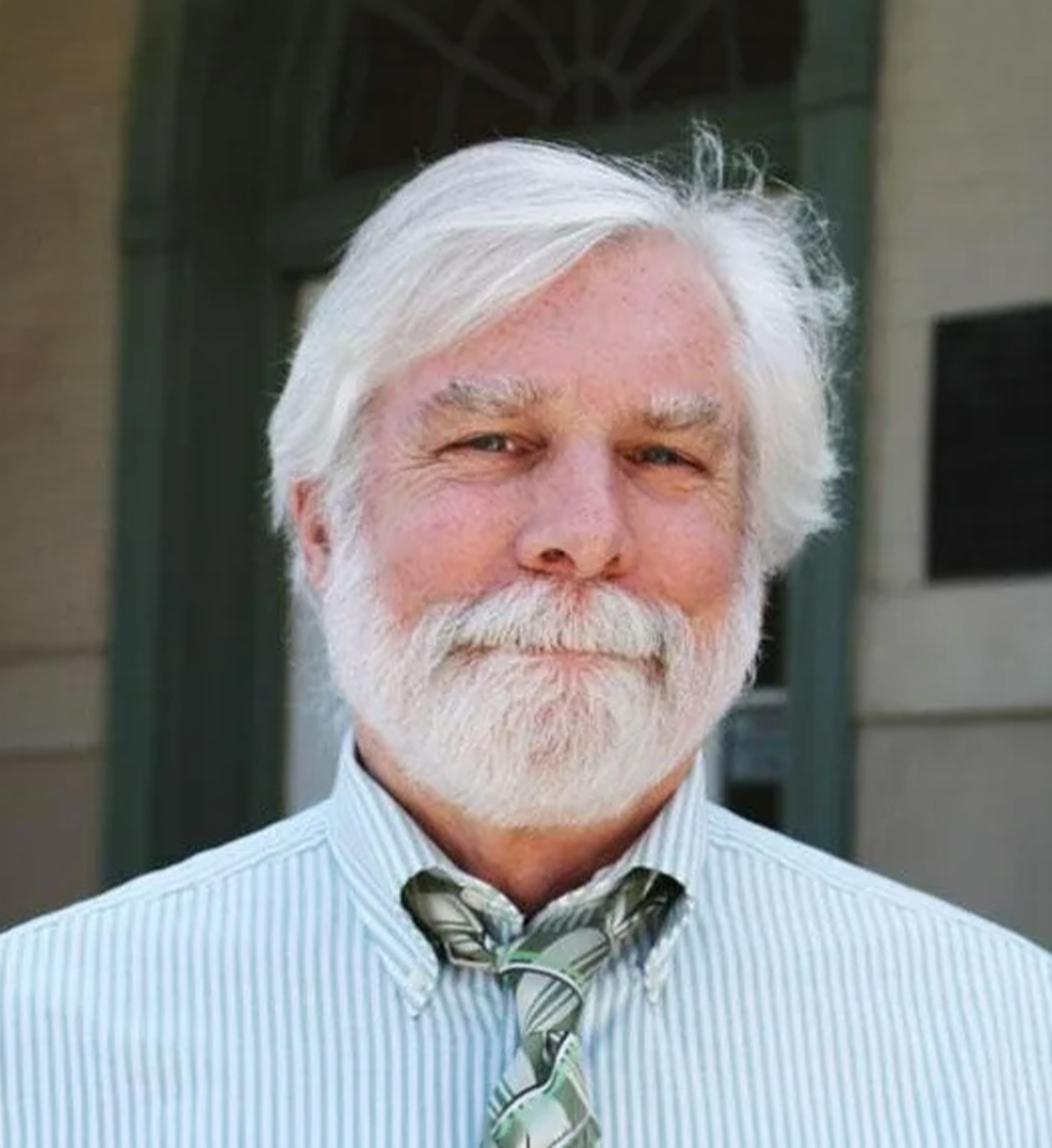 Dr. Michael smiling with white hair and a beard, wearing a striped dress shirt and patterned tie.